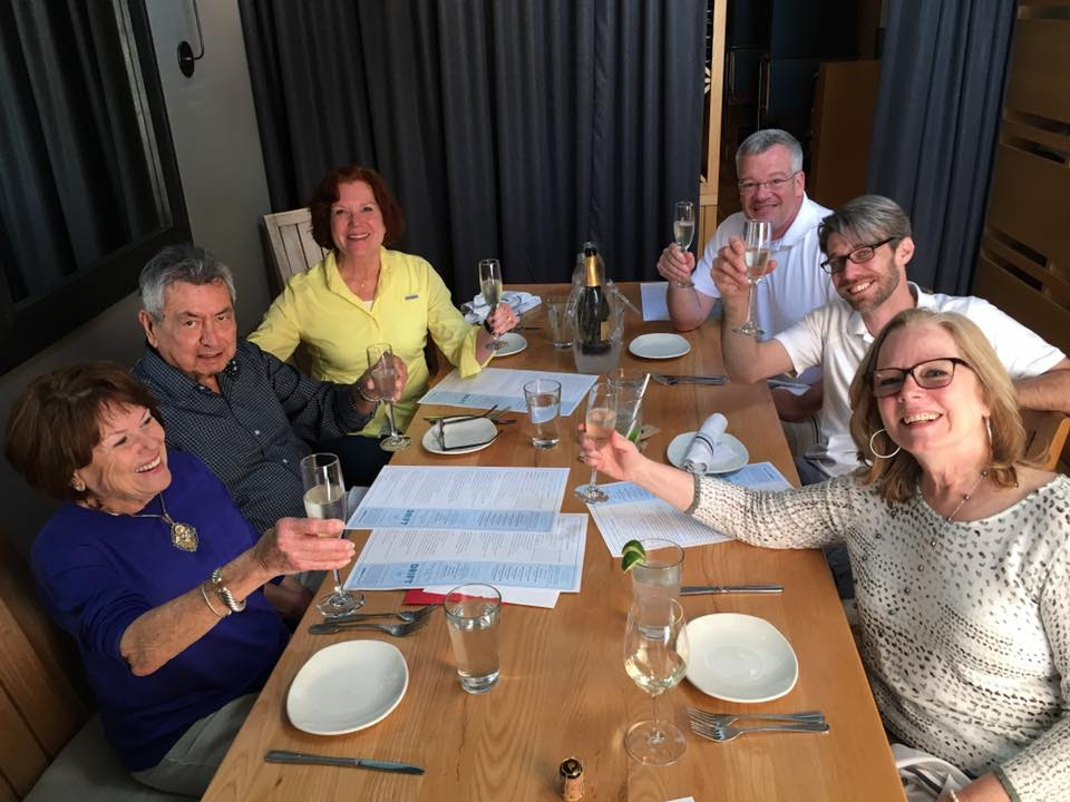 A group of six happy adults sitting around a wooden restaurant table, smiling and raising glasses in a cheerful toast during a meal gathering, representing community wellness and holistic living with My Vitality Project.